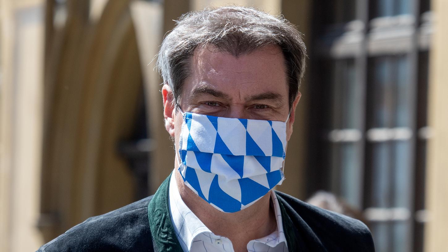 Markus Söder trägt einen Gesichtsschutz in den bayerischen Farben weiß und blau. Er hält strenge Auflagen in der Coronakrise weiter für gerechtfertigt.