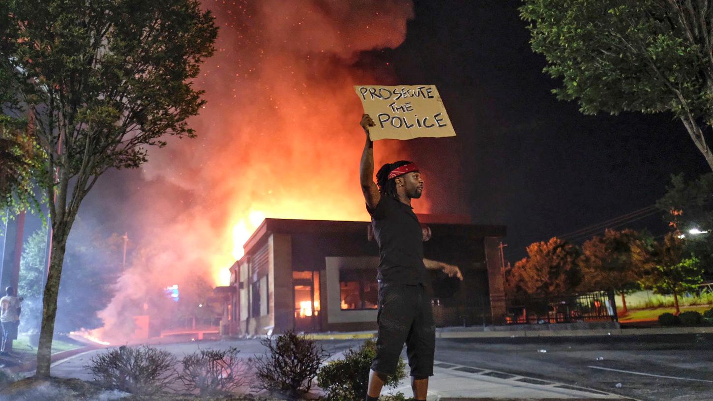 USA, Atlanta: Ein Mann hält ein Schild, während im Hintergrund ein Wendy's-Restaurant brennt. Ein 27-jähriger Afroamerikaner wurde am Freitagabend während eines Kampfes von der Polizei erschossen. Die Behörden untersuchen derzeit den Fall. 