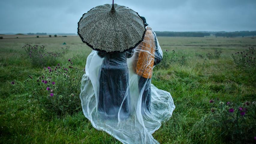 Menschen stehen neben dem aufgrund der Corona-Pandemie geschlossenen Bauwerk Stonehenge im Südwesten Englands und schützen sich mit Schirm und durchsichtiger Folie vor dem Regen - ausgerechnet während einer Versammlung zur Sommersonnenwende, um den Anbruch des längsten Tages im Jahr zu feiern.