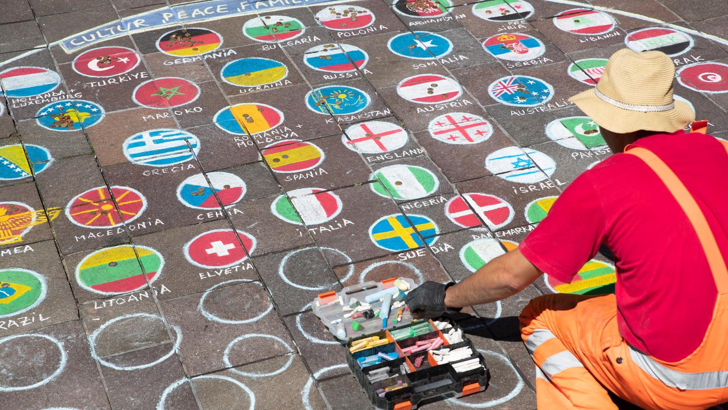 Ein Mann malt die Fahnen verschiedener Nationen auf das Pflaster der Fußgängerzone von Paderborn.