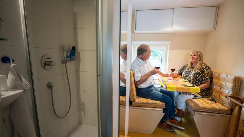Der gelernte Architekt Achim Schollenberger sitzt gemeinsam mit seiner Partnerin, der freien Journalistin Simone Stiefel im ersten Stockwerk ihres kleinen Wohnhauses am Tisch, der in eine Liegefläche verwandelt werden kann. Links ist das Bad mit Toilette und Duschkabine zu sehen.
