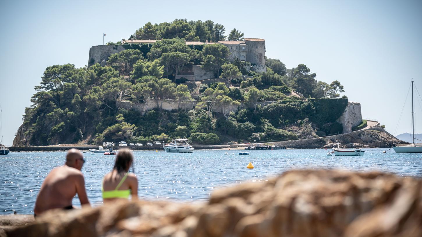 Badegäste sitzen im französischen Bormes-Les-Mimosas an einem Ufer gegenüber der früheren Festung "Fort de Bregancon" am Mittelmeer. 