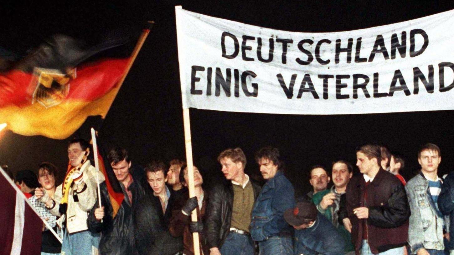 Mit der Deutschlandfahne und einem Transparent „Deutschland Einig Vaterland“ standen zahlreiche Berliner Ende Dezember 1989 auf der Berliner Mauer am Brandenburger Tor. 1990, im Jahr darauf, wurde dann die Deutsche Einheit besiegelt.