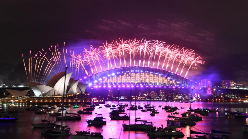 Im Hafen treibende Boote werden vom Feuerwerk beleuchtet, das von der Sydney Harbour Bridge und dem Opernhaus (l) aus gestartet wird. In diesem Jahr raten die Behörden den Einwohnern, sich das Feuerwerk aufgrund der Coronavirus-Pandemie im Fernsehen anzusehen.