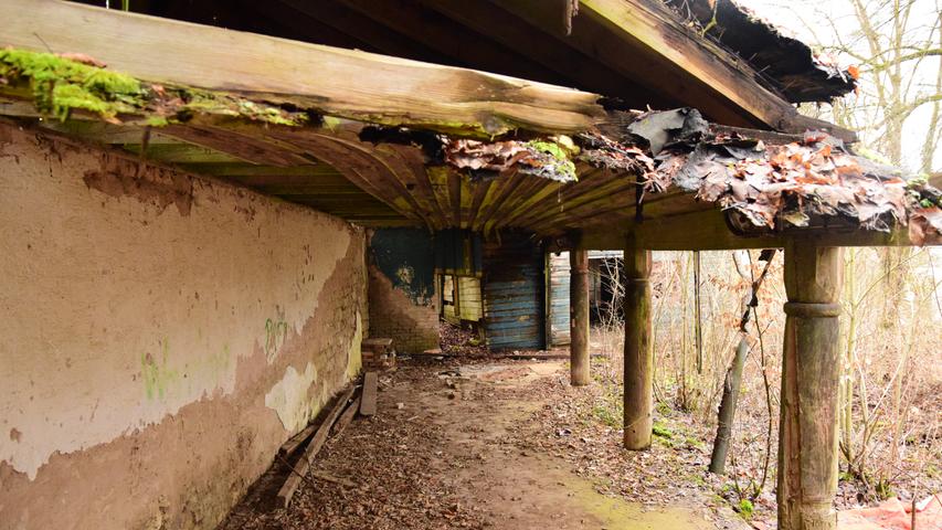 Dachbalken, die sich unter der Feuchtigkeit schon lange durchbiegen, gibt es auch entlang der überdachten Veranda auf der Ostseite des Kellers. Die Steinwand ist teilweise mit Graffiti besprüht. 