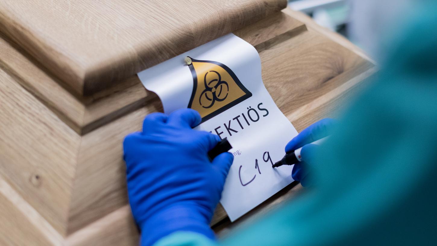 Ein Bestattungsunternehmer schreibt „C19“ auf ein Schild mit dem Hinweis „Infektiös“ auf einen Sarg mit einem Verstorbenen, der an oder mit dem Coronavirus gestorben ist. Der Leichnam darin darf nicht bekleidet sein, sondern wird in mit Desinfektionsmittel getränkte Leinentücher eingewickelt.
Ein Bestattungsunternehmer schreibt „C19“ auf ein Schild mit dem Hinweis „Infektiös“ auf einen Sarg mit einem Verstorbenen, der an oder mit dem Coronavirus gestorben ist. Der Leichnam darin darf nicht bekleidet sein, sondern wird in mit Desinfektionsmittel getränkte Leinentücher eingewickelt.