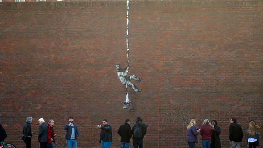 Menschen fotografieren ein Banksy-Werk, das einen ausbrechenden Häftling darstellt und an die Wand eines ehemaligen Gefängnisses gemalt wurde. Der improvisierte Strick, mit dem sich der Mann abseilt, ähnelt unten einem langen Stück Papier, an dessen Ende eine Schreibmaschine baumelt. 