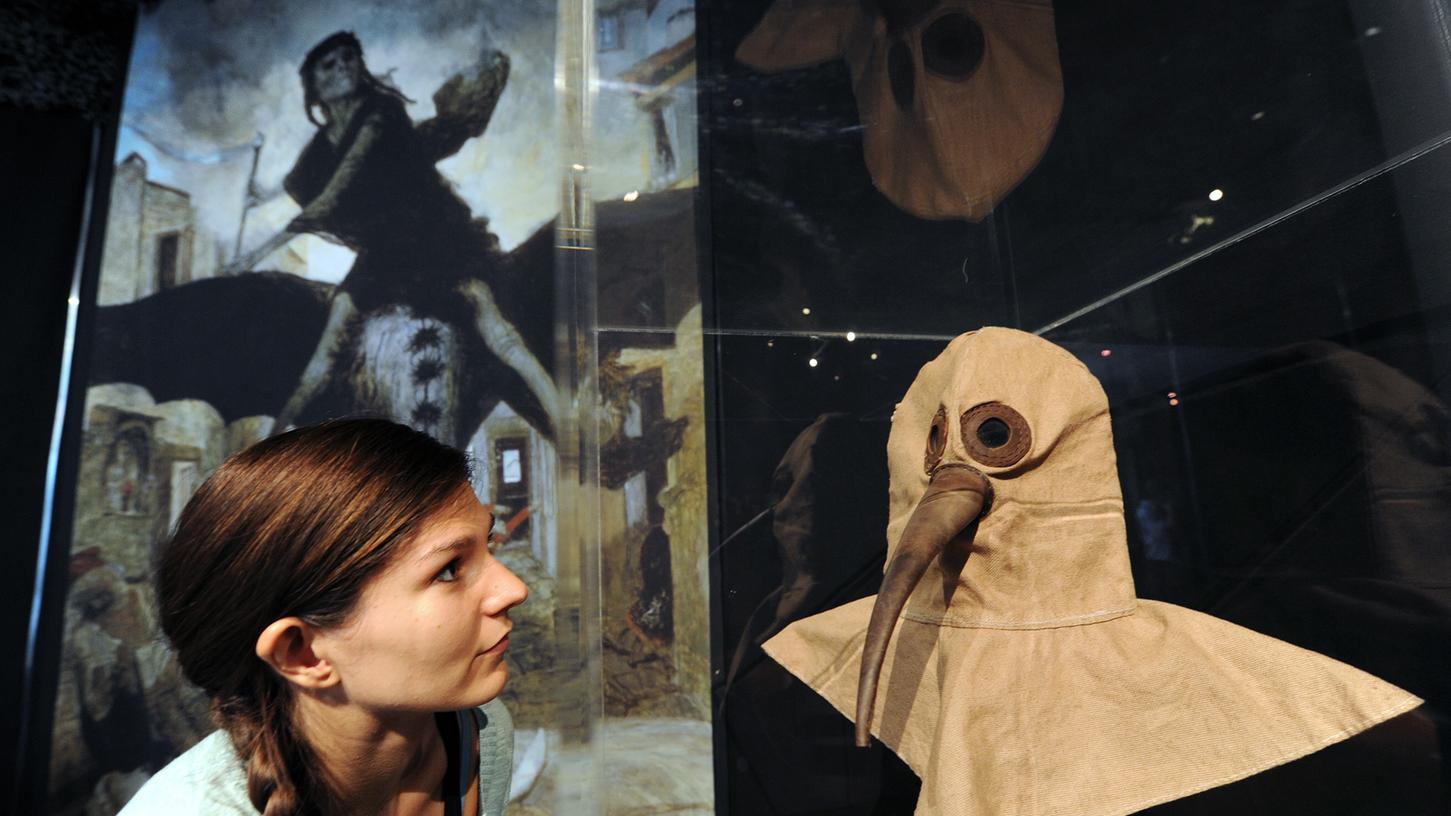 Maske, Quarantäne und Passierschein im 16. Jahrhundert: Eine Restauratorin betrachtet im Kasseler Museum für Sepulkralkultur (Totenkult) vor einer großformatigen Ansicht des „Schwarzen Todes“ (Pest) die Reproduktion einer Pestarzt-Maske.