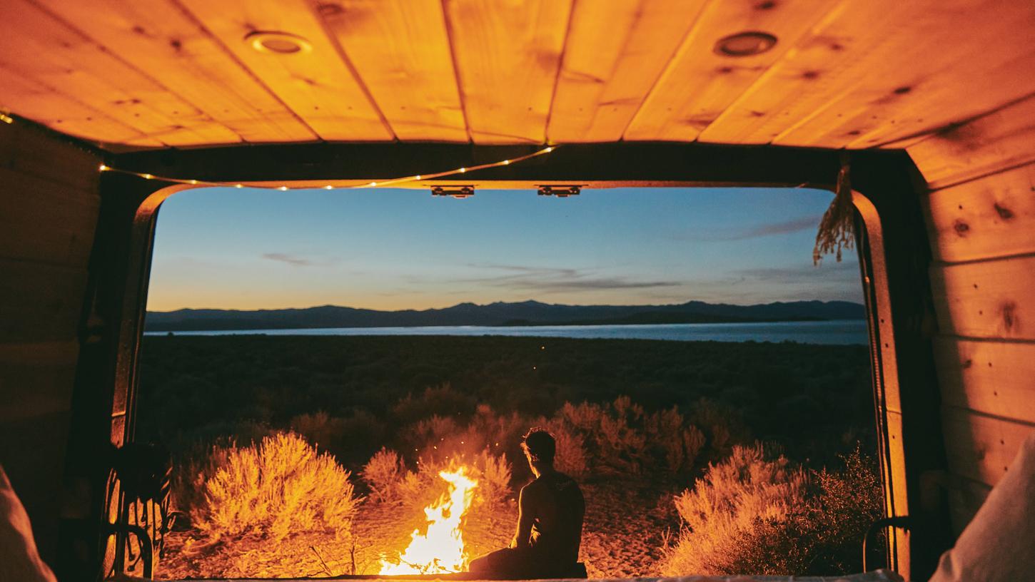 Ein Ideal des Mikroabenteuers: Ein Kleinbus, heimelig mit Holz verkleidet, davor ein Lagerfeuer und der Ausblick in den abendlichen Horizont. Reisefreiheit, die auch in Coronazeiten möglich ist.