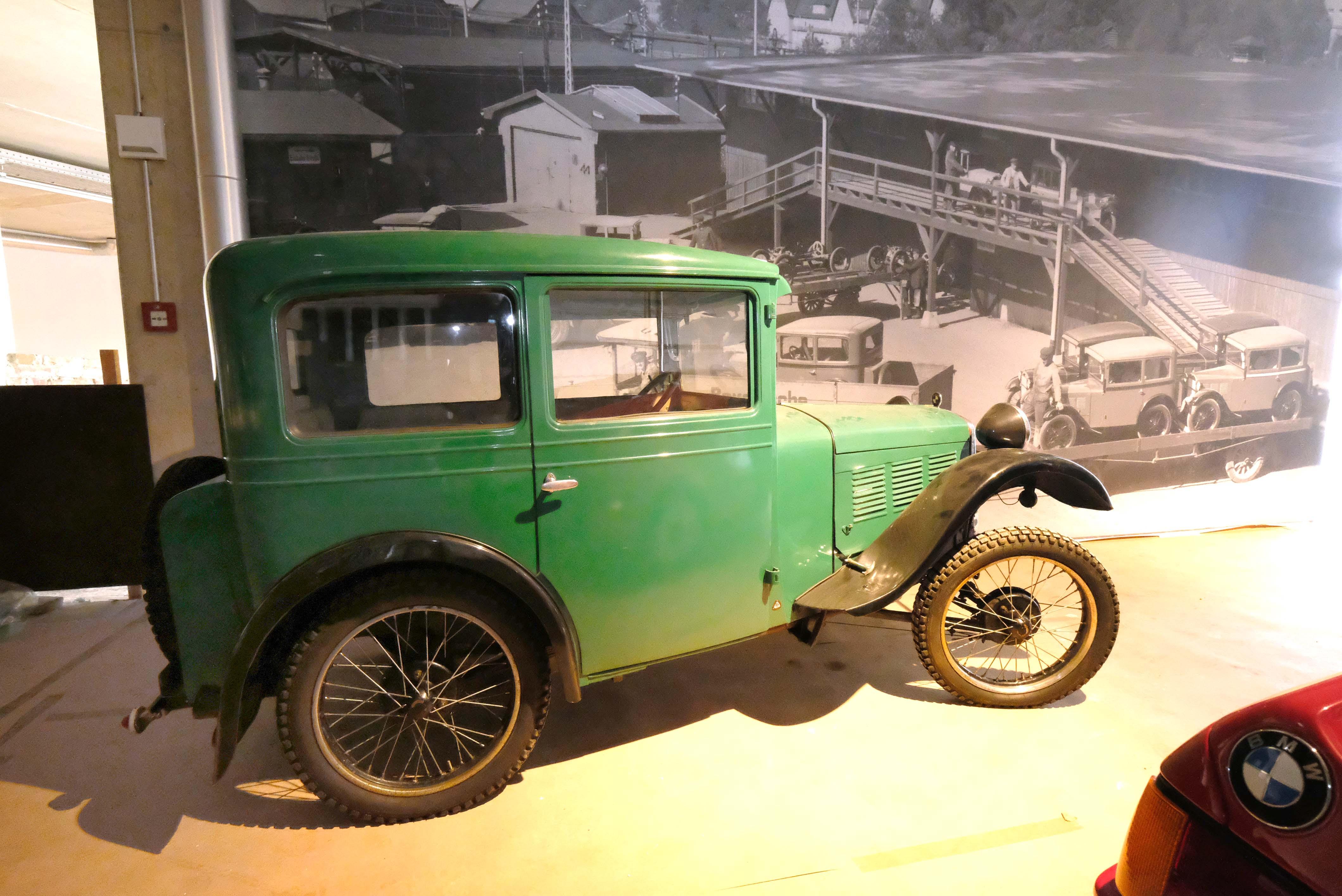 Im Hintergrund sieht man auf einer historischen Fotografie die Auslieferung im BMW-Werk in Eisenach. Davor steht ein Modell, wie es auf der Fotografie abgebildet ist.