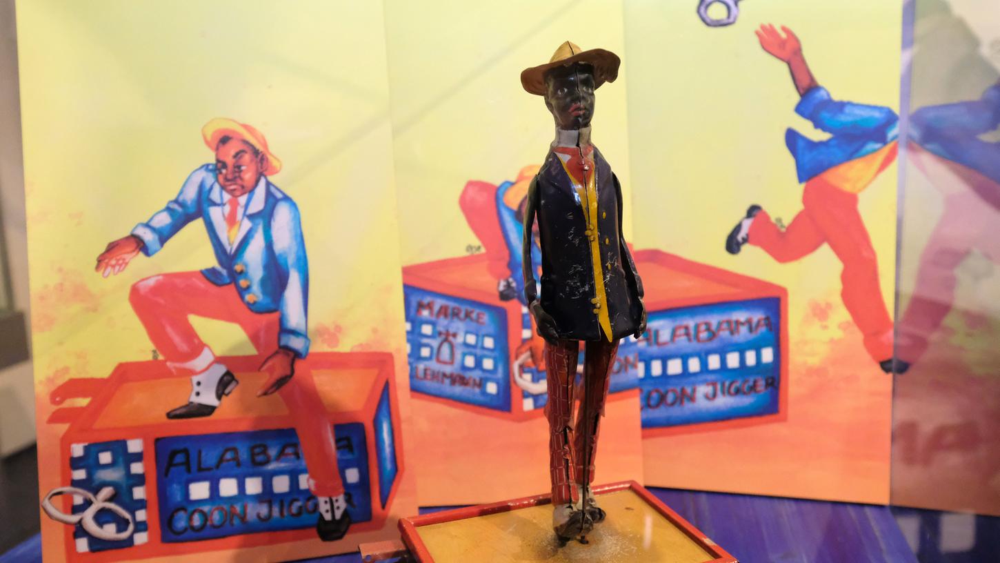 Die beanstandete Blechfigur von 1912 und die moderne Interpretation auf den plakativen Bildern im Hintergrund: Der Schwarze zieht den Schlüssel ab, der ihn zum Tanzen zwingt, wirft ihn weg und verlässt das Podium. Die beanstandete Blechfigur von 1912 und die moderne Interpretation auf den plakativen Bildern im Hintergrund: Der Schwarze zieht den Schlüssel ab, der ihn zum Tanzen zwingt, wirft ihn weg und verlässt das Podium.