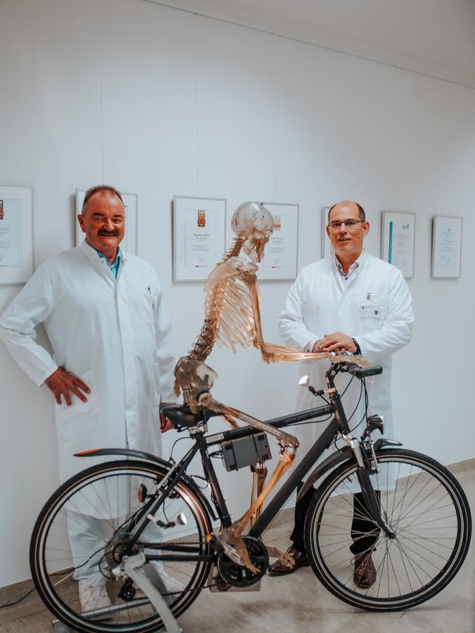 Orthopädie-Chefarzt Prof. Dr. Thomas Tischer (r.) und Oberarzt Dr. Torsten Elbracht mit einem Schaustück im Waldkrankenhaus St. Marien Erlangen, einem Skelett auf einem Fahrrad.