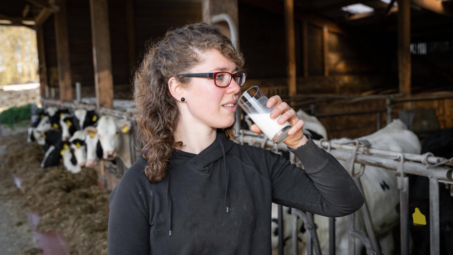  Eine junge Landwirtin trinkt ein Glas Milch vor einigen Kühen im Kuhstall. 

