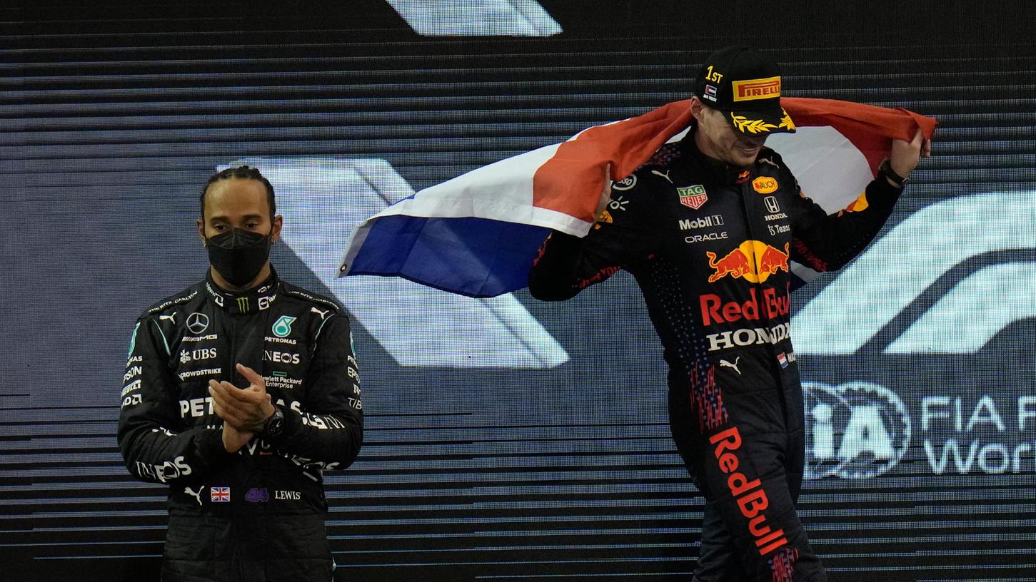 Max Verstappen jubelt mit der niederländischen Flagge über seinen Sieg, während neben ihm der Zweitplatzierte Lewis Hamilton bedröppelt schaut.
