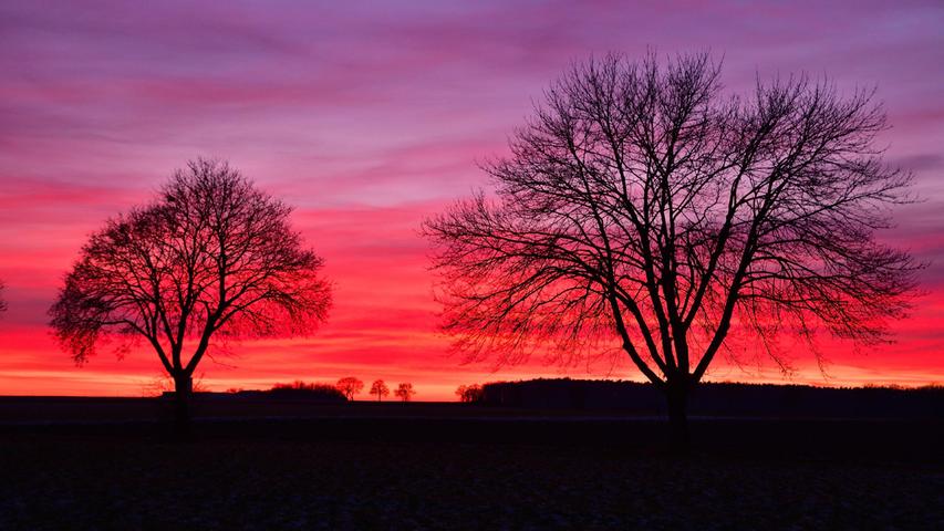 Die beiden Bäume heben den Kontrast zu dem stimmungsvollem Sonnenuntergang mit prächtigen Farben bei Erlangen schön hervor.
