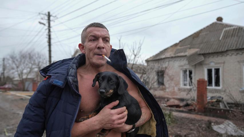 Ein Mann hält einen Hund, während er an einem nach russischem Beschuss beschädigten Haus vorbeigeht.
