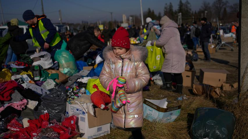 Ein ukrainisches Mädchen steht mit einem Spielzeug in der Hand neben einem Stapel gespendeter Kleidung am Grenzübergang. 