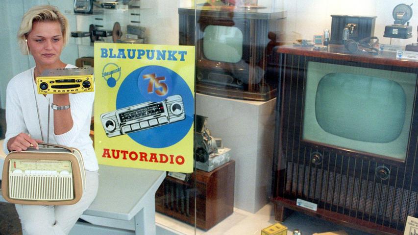 Vor Radio- und Fernseh-Geräten aus der 75-jährigen Blaupunkt-Produktion hält Unternehmens-Mitarbeiterin Anja Markworth 1998 in Hildesheim ein Kofferradio aus den 60er Jahren und ein Autoradio mit gelbem Outfit.