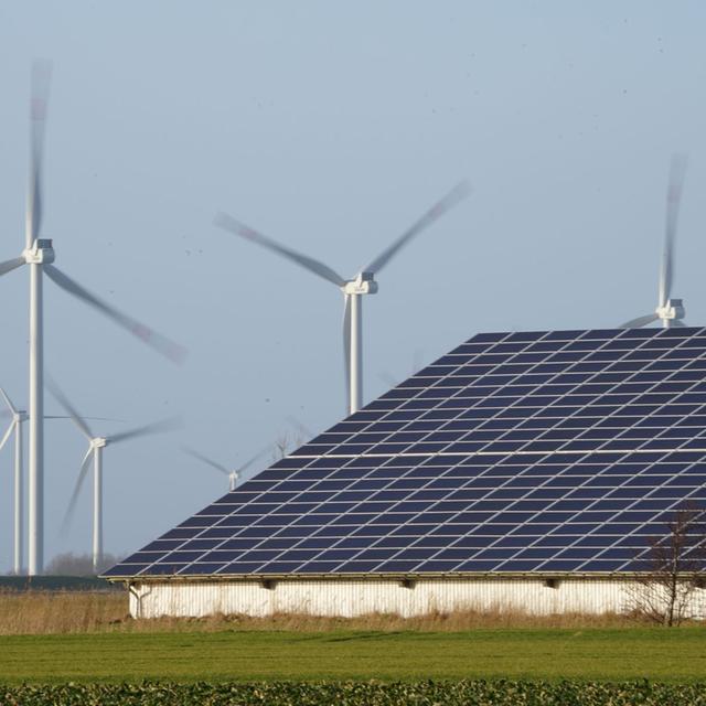 Windenergieanlagen stehen neben einer Halle mit Photovoltaik-Anlagen.