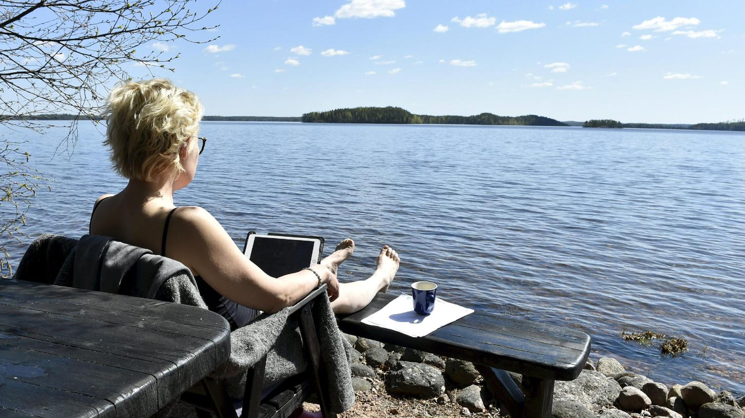 ARCHIV - 25.05.2020, Finnland, Kouvola: Eine Frau arbeitet am Ufer des Sees Vuohijärvi. Während der Corona-Pandemie wanderten viele Beschäftigten ins Home-Office. Aber mobil im Ausland arbeiten? Deutschlands große Unternehmen handhaben das ganz unterschiedlich. (zu dpa "Nach Home-Office: So funktioniert mobiles Arbeiten im Ausland") Foto: Jussi Nukari/Lehtikuva/dpa +++ dpa-Bildfunk +++ ARCHIV - 25.05.2020, Finnland, Kouvola: Eine Frau arbeitet am Ufer des Sees Vuohijärvi. Während der Corona-Pandemie wanderten viele Beschäftigten ins Home-Office. Aber mobil im Ausland arbeiten? Deutschlands große Unternehmen handhaben das ganz unterschiedlich. (zu dpa "Nach Home-Office: So funktioniert mobiles Arbeiten im Ausland") Foto: Jussi Nukari/Lehtikuva/dpa +++ dpa-Bildfunk +++