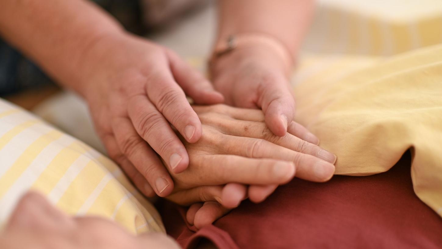 Eine Hospizmitarbeiterin hält die Hand eines todkranken Menschen, der in einem Hospiz im Bett liegt.