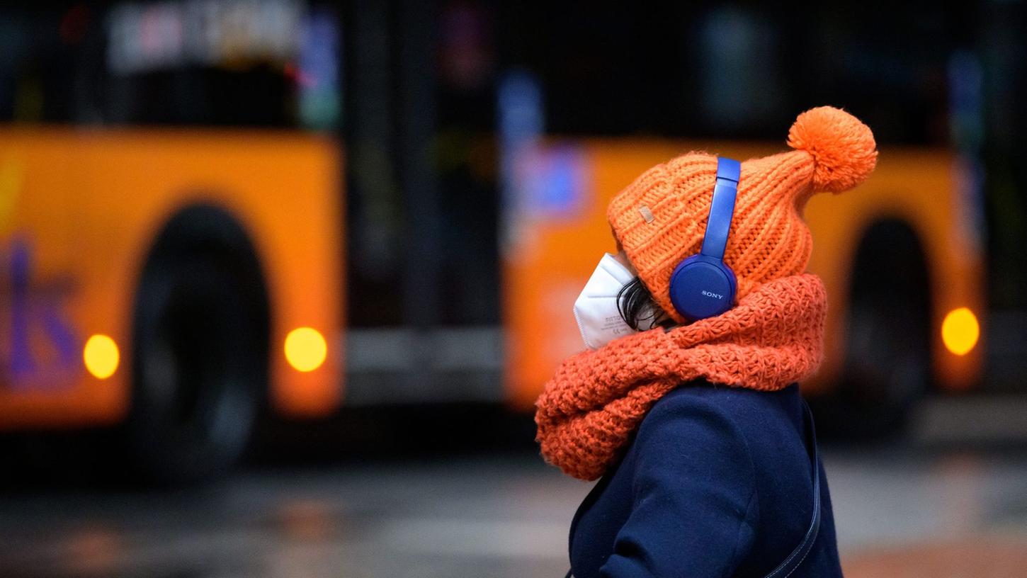 Eine Frau sitzt mit FFP2-Maske, dicker Mütze und Kopfhörern an einem Busbahnhof, während ein Linienbus vorbeifährt.