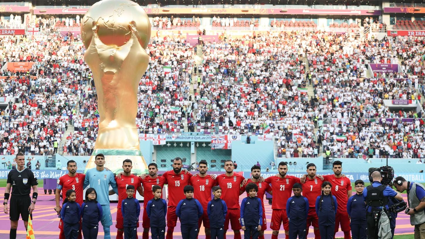 Spieler des Iran stehen beim Abspielen der Nationalhymne vor dem übergroßen Weltpokal im Stadion in Katar.