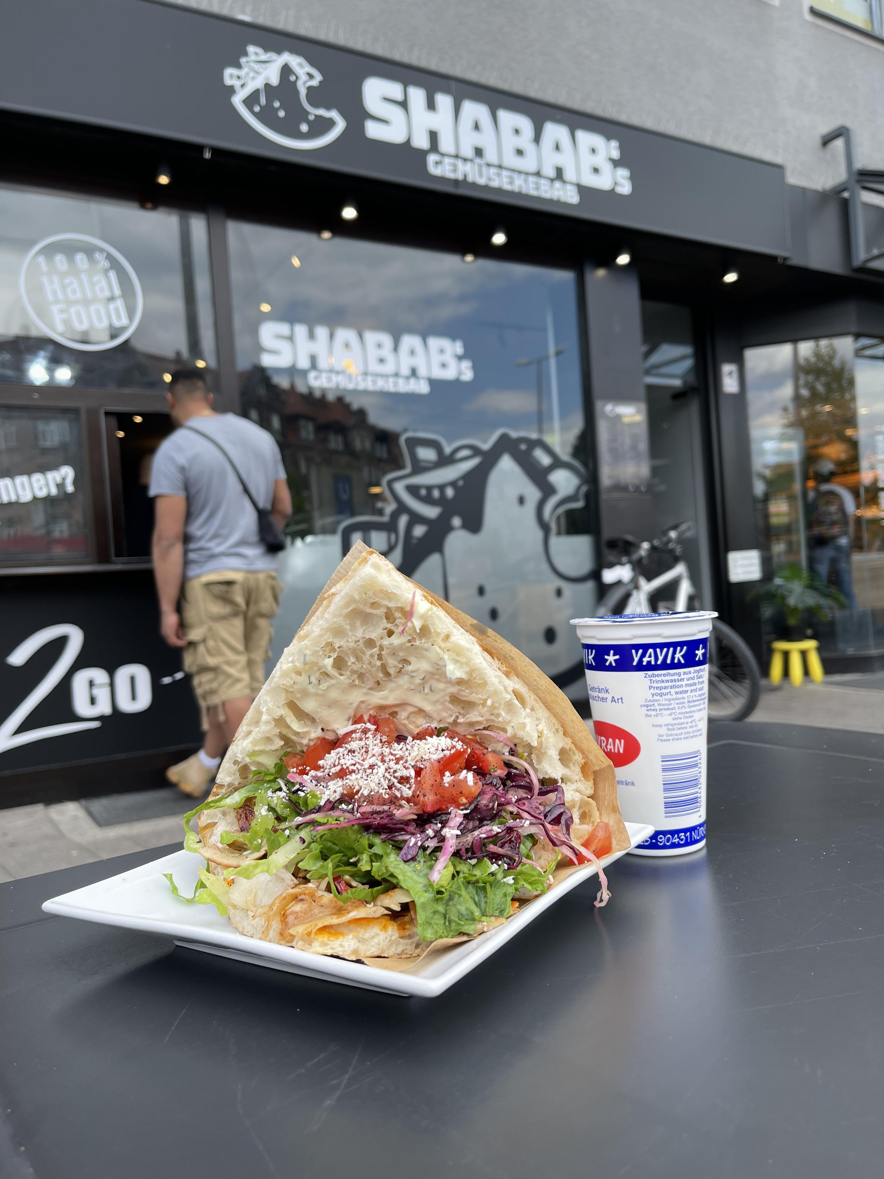 Am Friedrich-Ebert-Platz findet sich in der Bucher Straße 43 Shabab's Gemüsekebab. Und so sieht ein klassischer Kebab von Shabab's aus: knuspriges Fladenbrot, Fleisch, Romanasalat, Tomaten, Blaukraut, rote Zwiebeln, gebratenes Gemüse, etwa Zucchini, Paprika, Aubergine und Kartoffeln, Schafskäse, Sauce und ein paar Spritzer Zitrone zum Schluss. Die davon begeisterten Kundinnen und Kunden sorgen mit 199 Stimmen für Platz 2 im diesjährigen Döner-Voting.