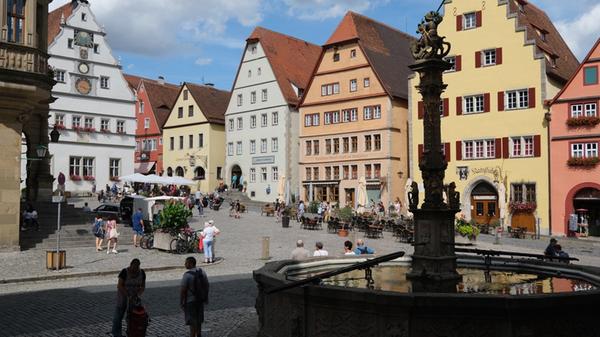 Es wird wohl niemanden geben, der Rothenburg ob der Tauber besucht und nicht am Marktplatz vorbeischaut. Aber häufig ist es der Blick auf das Rathaus, der im Mittelpunkt steht. In Wirklichkeit hat der zentrale Platz aber viele interessante Facetten, so wie diese Perspektive mit dem St. Georgsbrunnen (15. Jahrhundert) im Vordergrund auf die herrschaftlichen Häuser des reichsstädtischen Patriziats ihren Reiz hat. Hier versammeln sich viele Sehenswürdigkeiten. Links die Ratstrinkstube mit der großen Uhr, links und rechts davon kann man täglich das Glockenspiel vom Meistertrunk mit den Holzfiguren Tilly und Nusch bewundern. Und auf dem Marktplatz findet heutzutage wieder Leben statt, denn er ist teilweise gastronomisch bestuhlt.