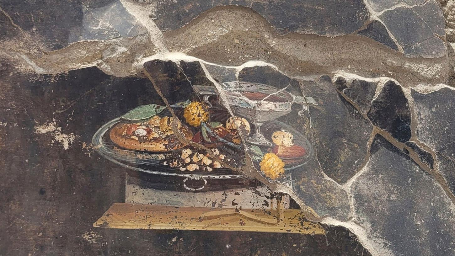 Die Wand eines pompejanischen Hauses mit einem Fresko, das einen Tisch mit Speisen darstellt, darunter auch eine Art Pizza.