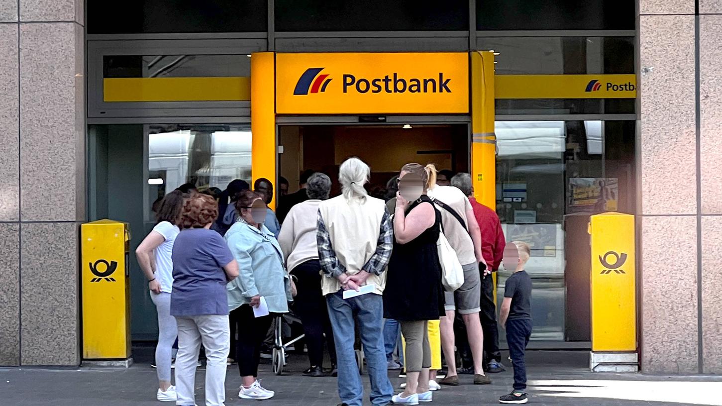 Kunden stehen in einer Warteschlange vor der Filiale einer Postbank. 
