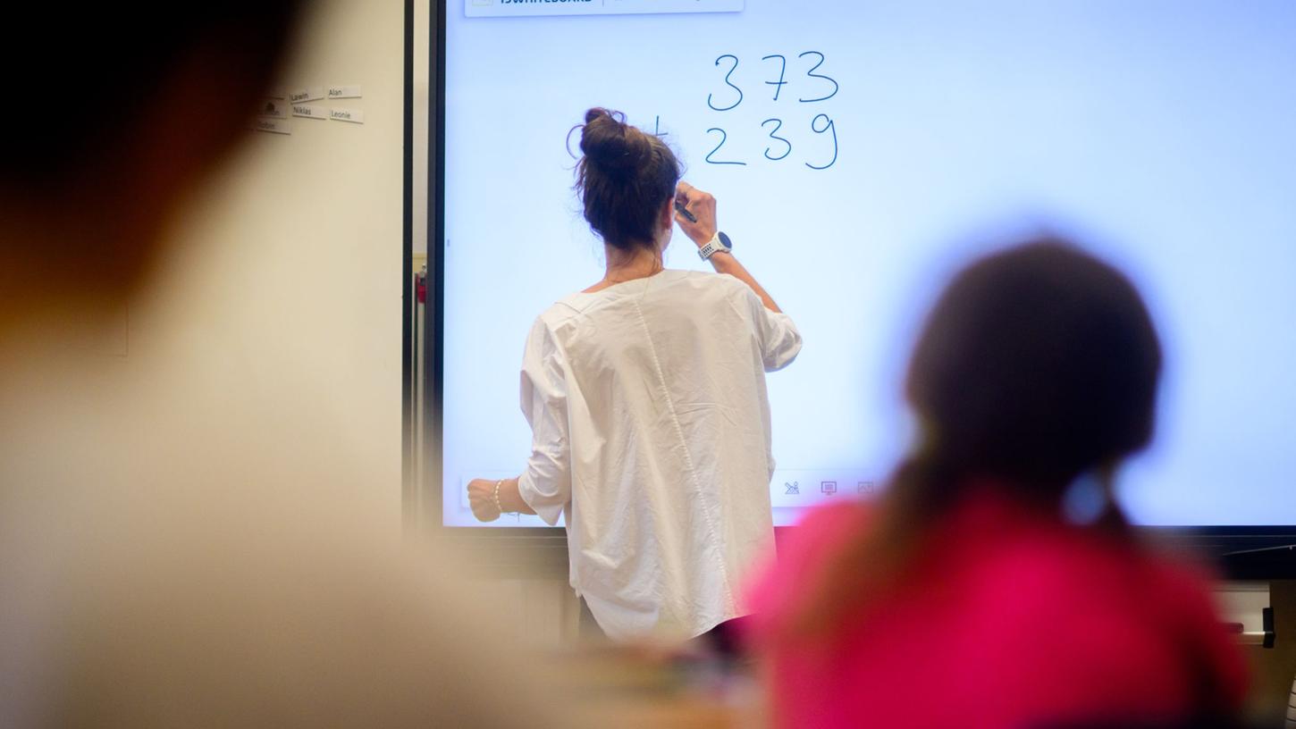 Eine Lehrerin schreibt eine Mathematikaufgabe auf eine digitale Schultafel im Klassenraum einer 4. Klasse einer Grundschule.