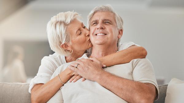 Happy senior couple, kiss and hug on sofa in living room for embrace, love or affection at home. Mature woman kissing and hugging man with smile in care, trust or support on lounge couch in house