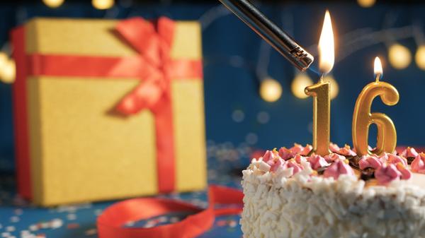 White birthday cake number 16 golden candles burning by lighter, blue background with lights and gift yellow box tied up with red ribbon. Close-up