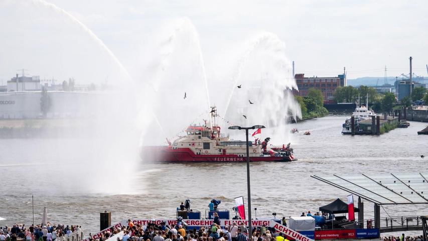 Ein Feuerlöschboot fährt an den Landungsbrücken mit Wasserfontänen entlang.