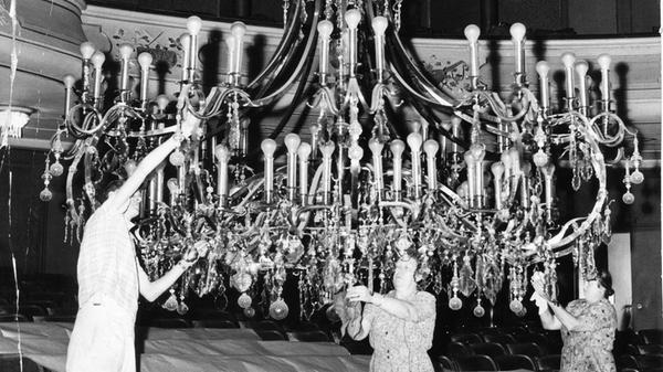 Putzaktion im Opernhaus Nürnberg im August 1959. Höhepunkt der alljährlichen Grundreinigung war das Herabsenken des riesigen Kronleuchters über dem Parkett. Per Hand musste der über 1.100 Kilogramm schwere und an starken Stahlseilen befestigte Lüster von mehreren Arbeitern ganz langsam bis hinunter zu den Parkettstühlen gesenkt werden. Tagelang wurden seine über 300 Kristallkugeln sowie rund 100 Glühlampen geputzt, poliert und von Elektrikern gewartet. Seit der von den Nationalsozialisten 1935 erfolgten Umgestaltung, der viele Jugendstil-Elemente zum Opfer fielen, erhellt der Lüster den Raum. Er hängt heute noch dort.