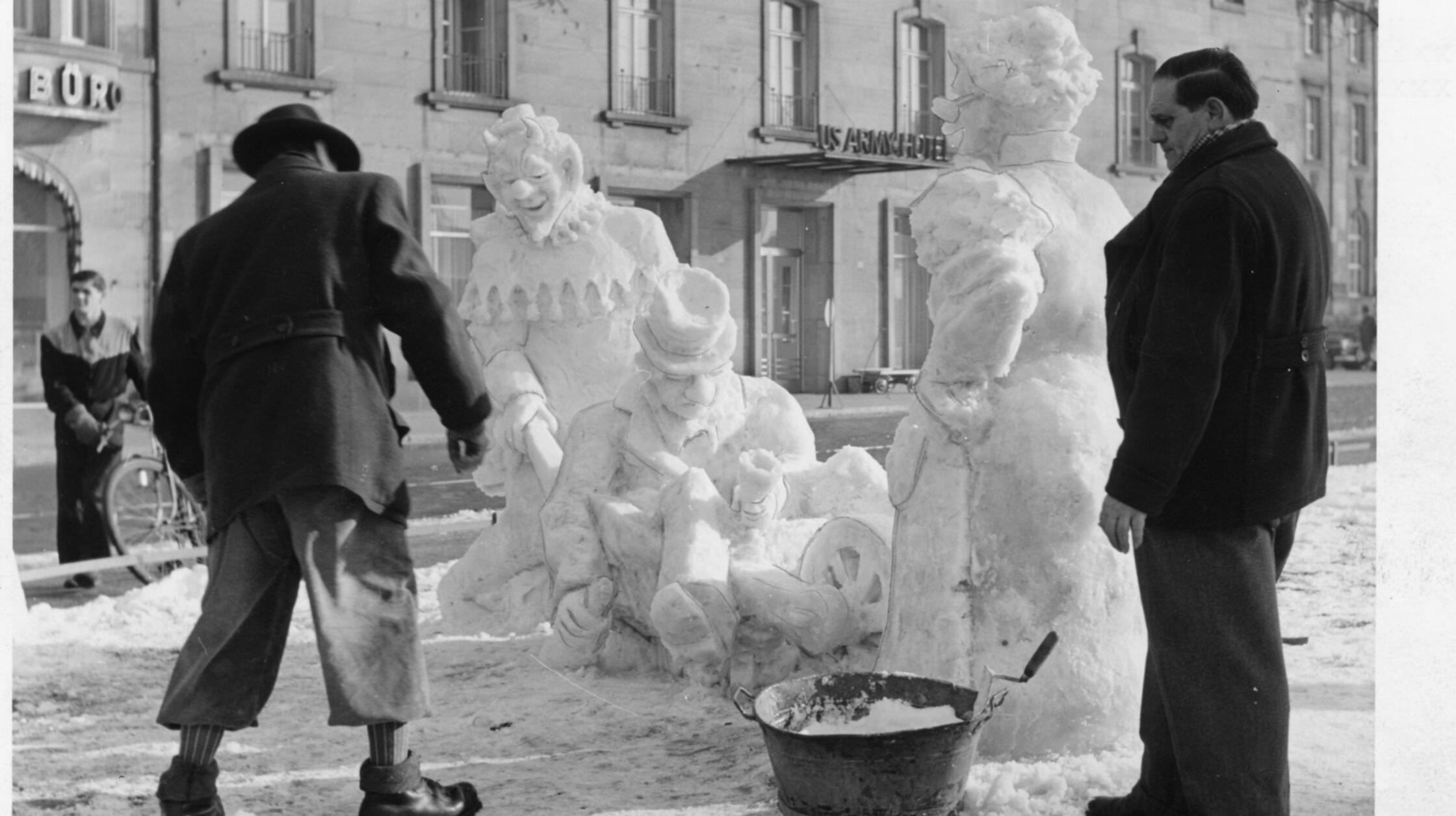 Eine humorvolle "Mahnung" an die Feiernden errichtete der Maler Konrad Seiler (hier in der modischen Knickerbocker-Hose links im Bild zu sehen) zum Fasching 1955 auf dem Bahnhofsplatz in Nürnberg. Seine eindrucksvolle Schneeskulptur zeigte einen Narren mit Teufelsmaske, welcher einen offenbar nach ausgiebigem Alkoholgenuss bewusstlosen Herren auf einer Schubkarre nachhause zu dessen wenig amüsierter Gattin brachte. Traditionell wurde in diesen Jahren regelmäßig zur fünften Jahreszeit ein entsprechend inspiriertes Kunstwerk dort erbaut, die damals noch schneereicheren Winter schufen die notwendige Voraussetzung dafür.