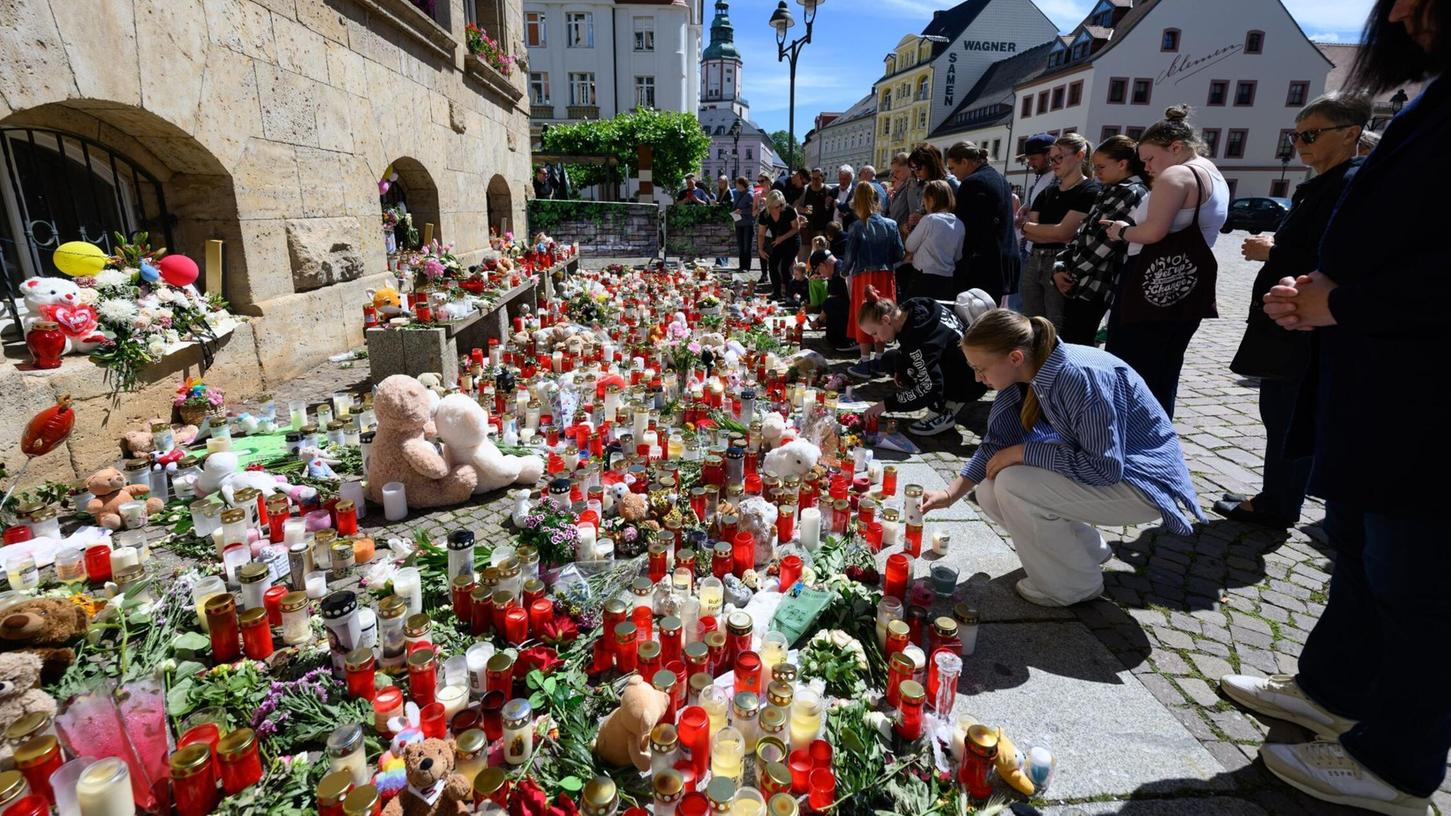 Menschen stehen am Rande eines Gottesdienstes für die getötete Valeriia in Döbeln vor Blumen und Kerzen.