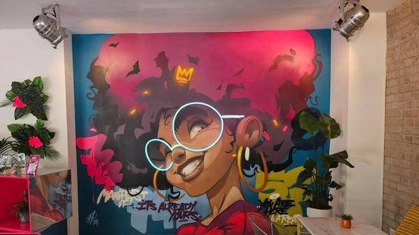 An der Rückwand des Restaurants prangt ein großes Graffiti. Der Künstler dahinter ist kein Geringerer als Hombre Suk. Ein Highlight - wortwörtlich - im Graffiti ist die leuchtende Brille, diese haben die Atlis selbst gestaltet und in das Kunstwerk eingebaut.