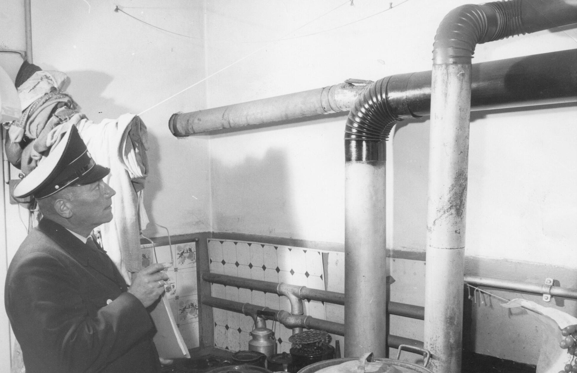 Feuerstättenschau zu Beginn der Heizperiode im Jahr 1959. Skeptisch betrachtet der Beamte eine eigenwillige Konstruktion aus Ofenrohren. Hinter Nürnbergs Türen gab es damals eine Vielfalt verschiedener Heizungen. Die Heiztechnik befand sich im Wandel. Ein mit Kacheln verkleideter Dauerbrandofen war zunehmend in vielen Wohnungen zu finden. Die Zentralheizung verbreitete sich, blieb aber noch den Betuchteren vorbehalten. Koks- und Kohleöfen waren rückläufig, Öl- und Gasheizung auf dem Vormarsch. Heute wiederum kehrt man sich von fossil betriebenen Heizanlagen ab, um sie mit klimafreundlicherer Technik zu ersetzen.
