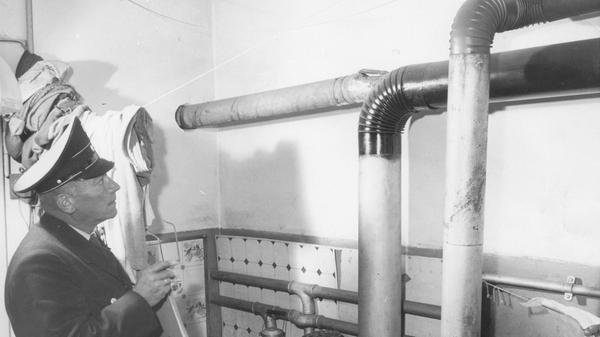 Feuerstättenschau zu Beginn der Heizperiode im Jahr 1959. Skeptisch betrachtet der Beamte eine eigenwillige Konstruktion aus Ofenrohren. Hinter Nürnbergs Türen gab es damals eine Vielfalt verschiedener Heizungen. Die Heiztechnik befand sich im Wandel. Ein mit Kacheln verkleideter Dauerbrandofen war zunehmend in vielen Wohnungen zu finden. Die Zentralheizung verbreitete sich, blieb aber noch den Betuchteren vorbehalten. Koks- und Kohleöfen waren rückläufig, Öl- und Gasheizung auf dem Vormarsch. Heute wiederum kehrt man sich von fossil betriebenen Heizanlagen ab, um sie mit klimafreundlicherer Technik zu ersetzen.