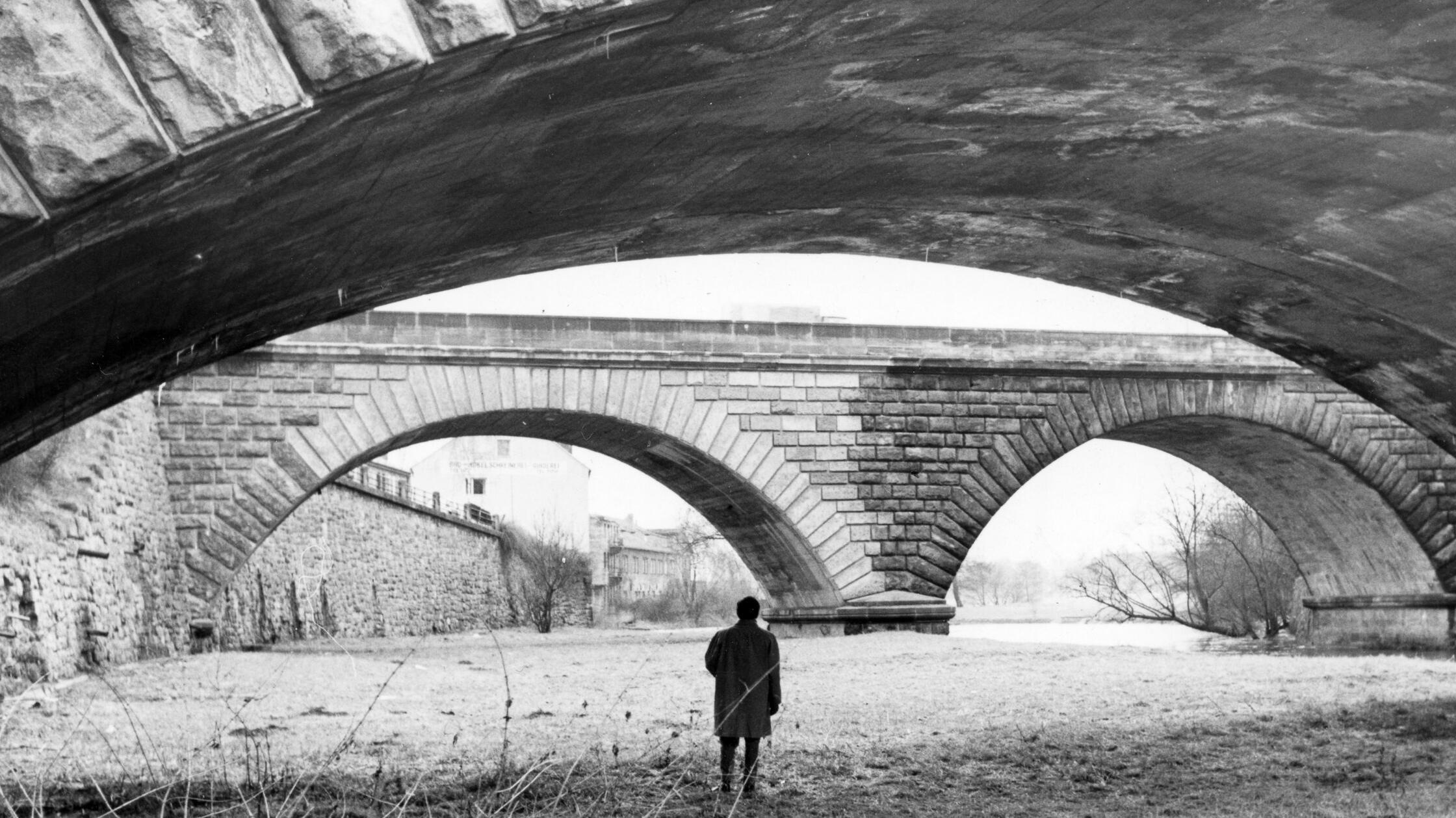An der Stadtgrenze zwischen Nürnberg und Fürth steht im Februar 1962 ein Mann im Flussbett und blickt auf die Bögen des Dooser Brückkanals. Eine malerische Umgebung. Doch aufgrund der Verkehrsplanung verschwand sie kurz nach der Entstehung dieser Aufnahme. Noch im Lauf der 60er Jahre wurden beide abgebildeten Brücken abgerissen und eine Schnellstraße in die Trasse des alten Ludwig-Donau-Main-Kanals gebaut. Stünde man heute an dieser Stelle, so wäre man direkt unterhalb der Kurgartenbrücke mitten auf dem Frankenschnellweg. Bis heute ein umstrittenes Verkehrsprojekt, dessen weiterer Ausbau kontrovers diskutiert wird.