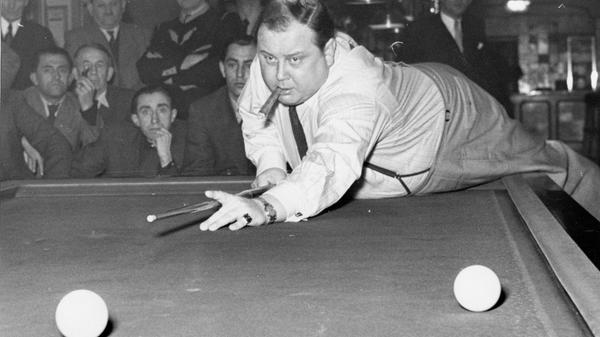 Zu Beginn der 1950er Jahre hatte Billard noch einen höheren sportlichen Stellenwert als heute. So richtete der 1. Billard-Club Nürnberg damals im Hotel Kaiserhof alljährlich Veranstaltungen aus, bei denen lokale und auch internationale Meister vor gespanntem Publikum gegeneinander antraten und ihr Können zeigten. Die Sportmode folgte dabei ihren eigenen Regeln, die Athleten erschienen klassisch-elegant. Im Eifer des Gefechtes war es aber wohl legitim, sich auch einmal des Sakkos zu entledigen. So wie es dieser Teilnehmer tat, der im Wettkampf vollen Körpereinsatz zeigte, jedoch auch eine gute Zigarre zu schätzen wusste.