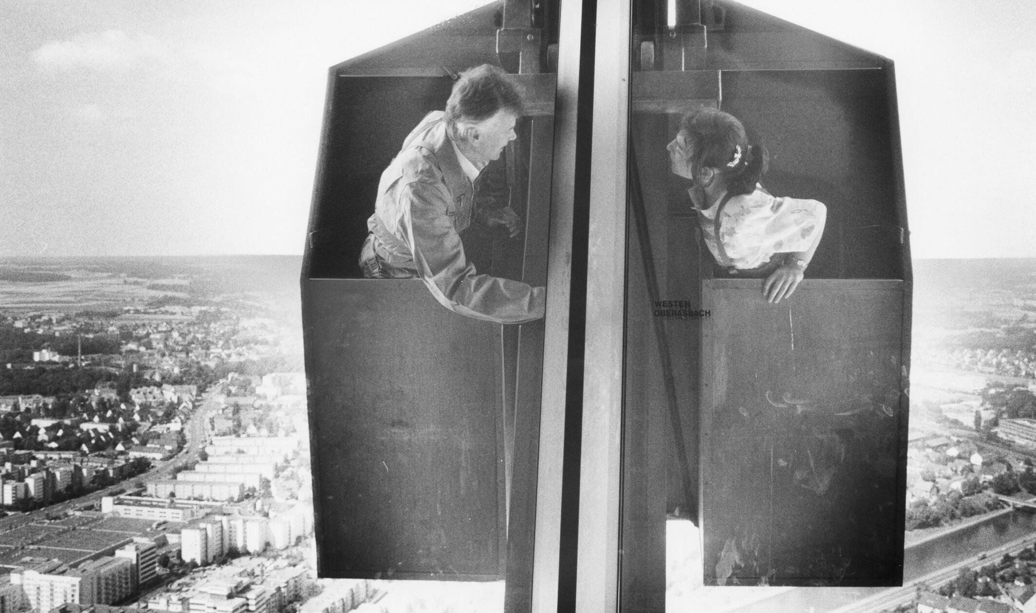 TÜV-Gutachter inspizierten im Juli 1990 den Außenaufzug für Fensterputzer des damals im Nürnberger Fernmeldeturm beheimateten Restaurants. Der Ausblick der Besucher des in 189 Meter Höhe gelegenen Lokals war schon zwei Jahre lang getrübt gewesen. Der TÜV hatte verboten, die Scheiben zu putzen, nachdem der Außenaufzug 1988 bei einem Sturm beschädigt worden war. Ursprünglich sollte der gesamte Pylon abgeseilt und am Boden geprüft werden. Dieses teure Manöver ersparte TÜV-Ingenieur Günther Bäuerlein (li.) der Stadt: Er vertraute auf seine Schwindelfreiheit und reparierte den Aufzug deutlich kostengünstiger in luftiger Höhe.