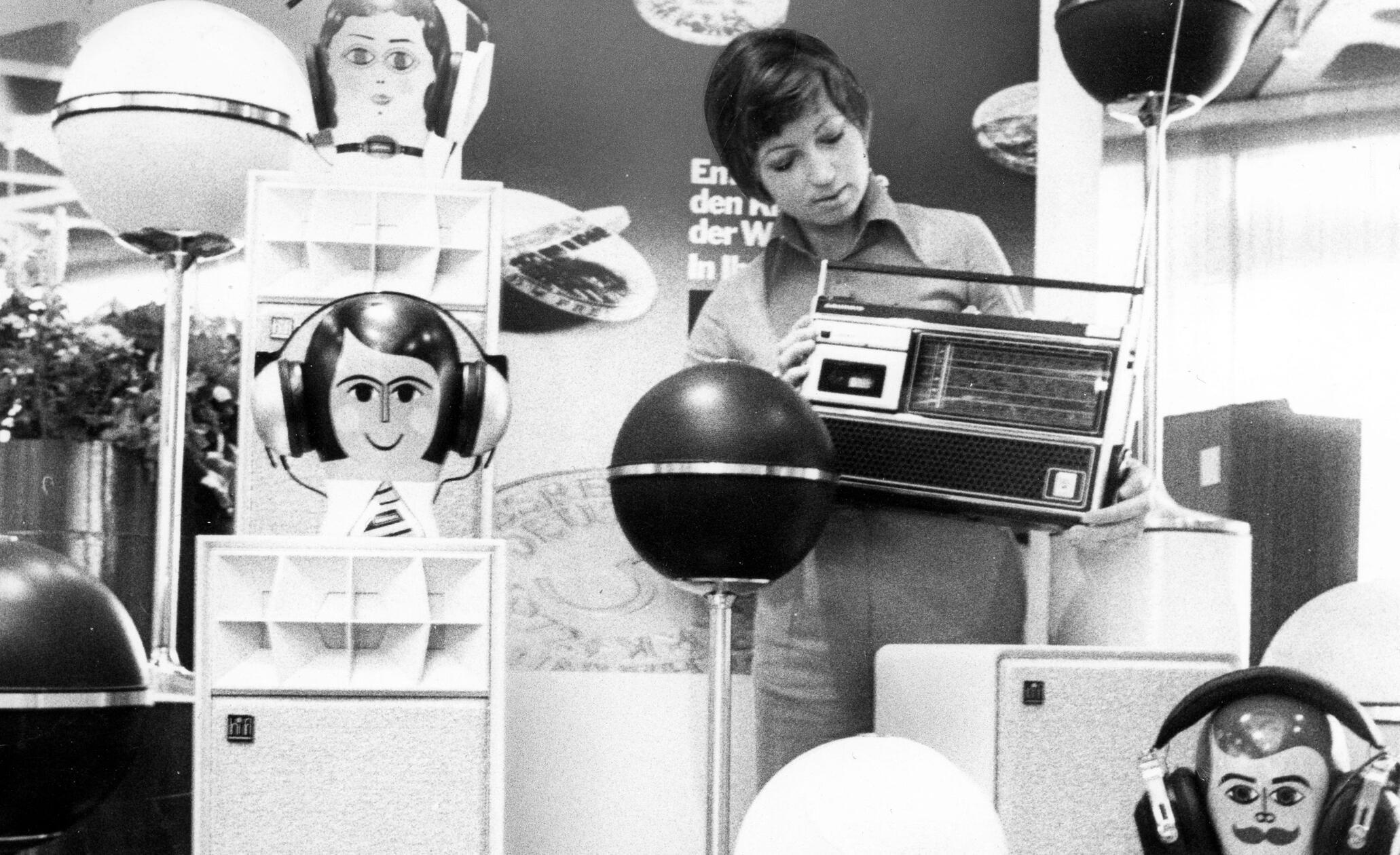 Anrührend retrofuturistisch mutet dieses Ensemble von Kugellautsprechern und Kopfhörern an, welches auf der von Grundig durchgeführten „Fachschau ‘74“ zu sehen war. Entsprechende Schauen fanden damals in 35 Städten statt, die HiFi-Branche erlebte in diesem Jahr, auch befördert durch den Weltmeisterschafts-Boom, einen spürbaren Aufschwung. Das Design der Elektronikprodukte folgte dabei konsequent dem Stil der damaligen Zeit. Die Dame mit dem Kassettenrekorder auf unserem Bild fügte sich mit ihrer modischen Frisur erstaunlich gut zwischen den durchaus eigenwillig gestalteten Kopfhörerständern ein.
