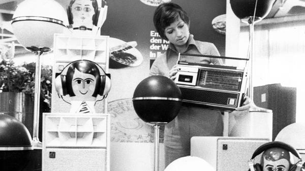 Anrührend retrofuturistisch mutet dieses Ensemble von Kugellautsprechern und Kopfhörern an, welches auf der von Grundig durchgeführten „Fachschau ‘74“ zu sehen war. Entsprechende Schauen fanden damals in 35 Städten statt, die HiFi-Branche erlebte in diesem Jahr, auch befördert durch den Weltmeisterschafts-Boom, einen spürbaren Aufschwung. Das Design der Elektronikprodukte folgte dabei konsequent dem Stil der damaligen Zeit. Die Dame mit dem Kassettenrekorder auf unserem Bild fügte sich mit ihrer modischen Frisur erstaunlich gut zwischen den durchaus eigenwillig gestalteten Kopfhörerständern ein.
