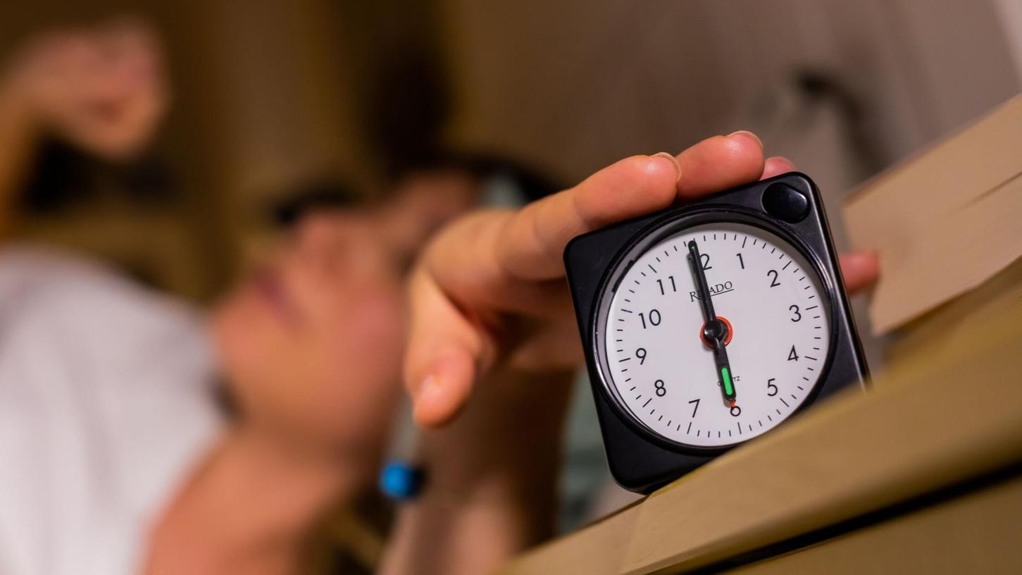 Eine Frau schaltet vom Bett aus einen auf sechs Uhr gestellten Wecker aus. Eine Frau schaltet vom Bett aus einen auf sechs Uhr gestellten Wecker aus.