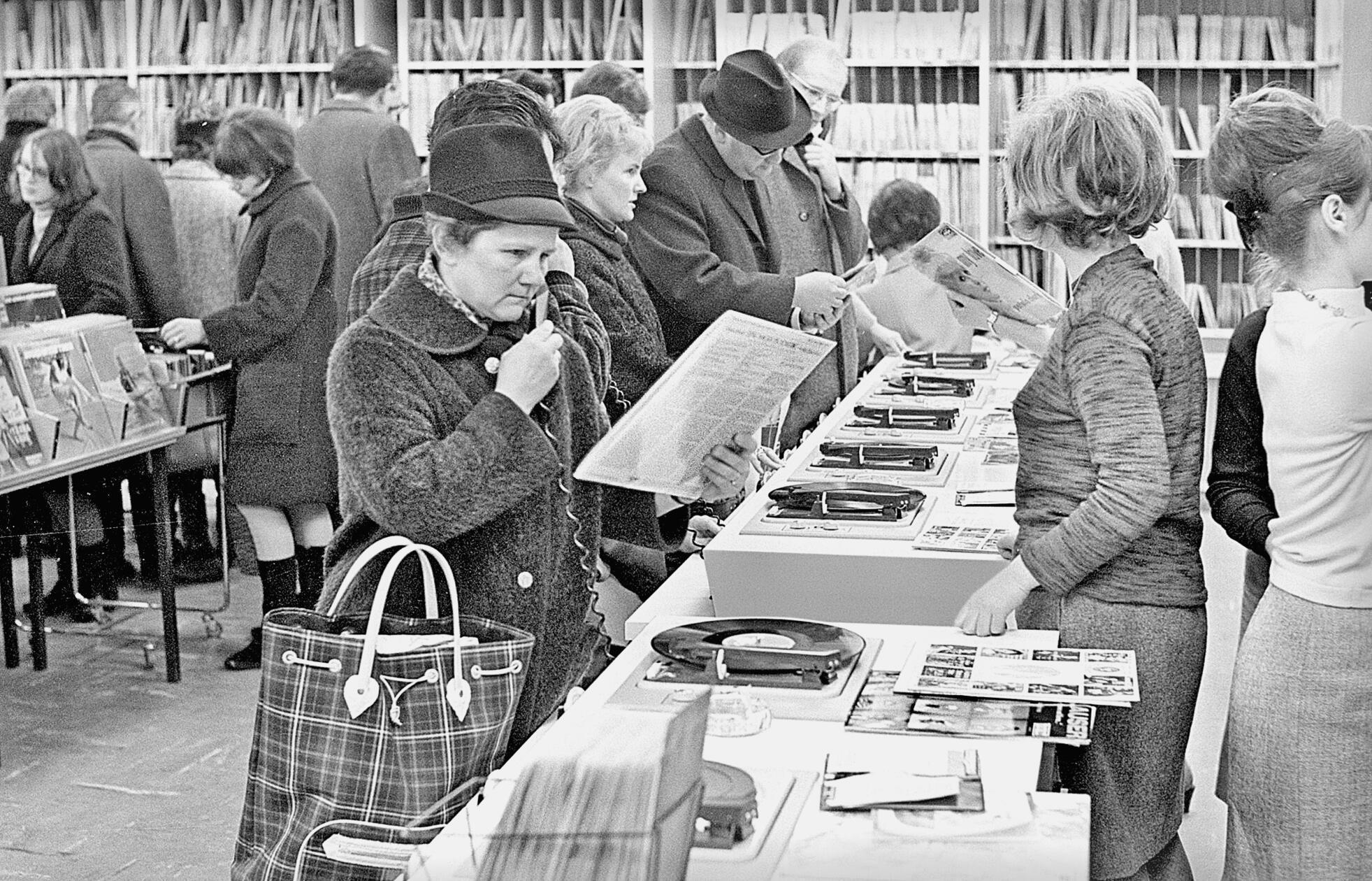 In Zeiten digitaler Musikplattformen mag es anachronistisch erscheinen, in den wenigen verbliebenen Plattenläden ist es jedoch auch heute oft noch möglich, vor dem Kauf in das Objekt der Begierde hineinzuhören. Genau so war es auch schon im Jahre 1966, mit dem Unterschied allerdings, dass sich dieses Angebot damals noch an die breite Bevölkerung und nicht nur an die selten gewordenen Liebhaber analoger Tonträger richtete. Statt eines Stereokopfhörers stand für die Hörprobe damals eine Art Telefonhörer ohne Sprechmuschel zur Verfügung, die Dame auf unserem Foto scheint gegenüber dem gebotenen Klangerlebnis allerdings etwas skeptisch gewesen zu sein.