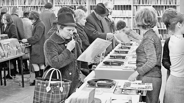 In Zeiten digitaler Musikplattformen mag es anachronistisch erscheinen, in den wenigen verbliebenen Plattenläden ist es jedoch auch heute oft noch möglich, vor dem Kauf in das Objekt der Begierde hineinzuhören. Genau so war es auch schon im Jahre 1966, mit dem Unterschied allerdings, dass sich dieses Angebot damals noch an die breite Bevölkerung und nicht nur an die selten gewordenen Liebhaber analoger Tonträger richtete. Statt eines Stereokopfhörers stand für die Hörprobe damals eine Art Telefonhörer ohne Sprechmuschel zur Verfügung, die Dame auf unserem Foto scheint gegenüber dem gebotenen Klangerlebnis allerdings etwas skeptisch gewesen zu sein.