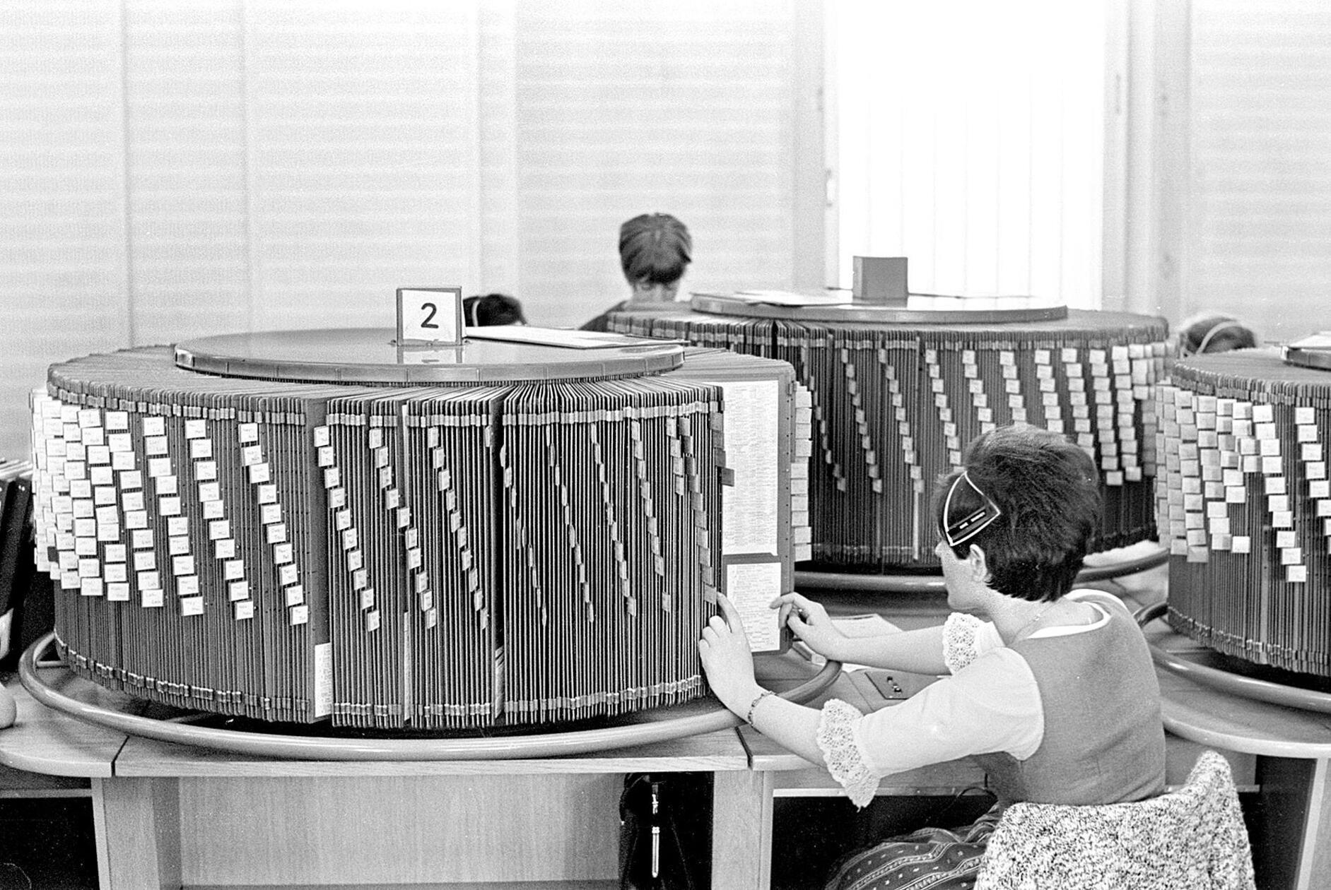 Wer bereits Mitte der 1960er Jahre glücklicher Besitzer eines Telefons war, und eine Telefonnummer benötigte, landete unter Umständen bei dieser hilfsbereiten Telefonistin an ihrem Arbeitsplatz im Fernmeldeamt 1 in der Karolinenstraße in Nürnberg. 8000 Anfragen gingen seinerzeit werktäglich bei der Telefonauskunft ein. In kürzester Zeit galt es – quasi im Handumdrehen – die gesuchte Telefonnummer in der sogenannten Drehauskunft zu finden, die die einzelnen Seiten des damaligen Amtlichen Fernsprechbuches enthielt. Um unvermeidliche Wartezeiten zu verkürzen, wurde deshalb 1967 auf Mikrofilmtechnik umgestellt.